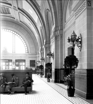 Three people sit on a wooden bench in a building with high domed ceilings, tiled floors, ornate lamps along the walls, and sunlight streaming in through a large arched window. 
